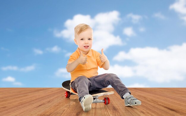 concetto di infanzia, sport, tempo libero, gesto e persone - ragazzino felice seduto sullo skateboard e mostrando i pollici in su sopra il cielo blu e lo sfondo del pavimento in legno