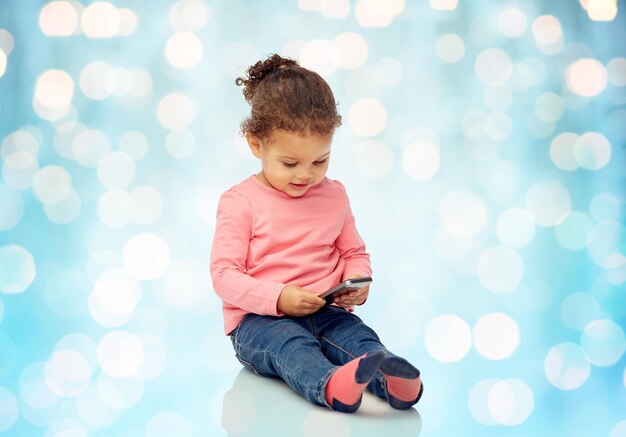 concetto di infanzia, tecnologia e persone - piccola bambina afroamericana sorridente che gioca con lo smartphone e si siede sul pavimento sopra lo sfondo blu delle luci delle vacanze