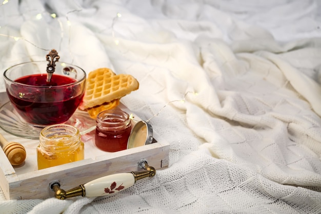 Concetto di inverno con coperta a maglia e tè caldo con waffer, marmellata, miele sul vassoio in legno