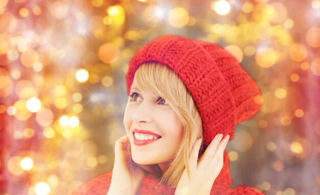 concetto di inverno, persone, natale e festività - donna sorridente felice con cappello rosso e sciarpa su sfondo di luci