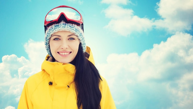 concetto di inverno, tempo libero, sport e persone - giovane donna felice in occhiali da sci su sfondo blu cielo e nuvole