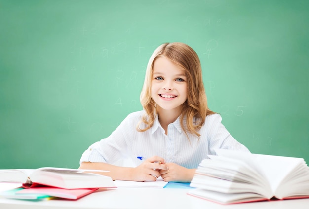 concetto di istruzione, persone, bambini e scuola - ragazza felice dello studente che si siede al tavolo con i libri e che scrive nel taccuino sopra il fondo del bordo di gesso verde