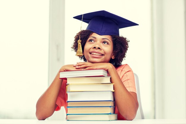 concetto di istruzione, scuola, conoscenza e persone - ragazza studentessa afroamericana sorridente felice in cappello da scapolo con libri seduti a tavola e sognando a casa