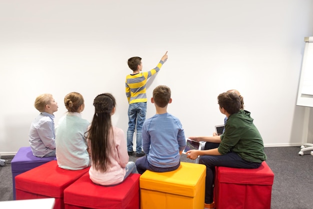 concetto di istruzione, scuola elementare, apprendimento e persone - ragazzo studente con gli amici che mostrano al muro bianco