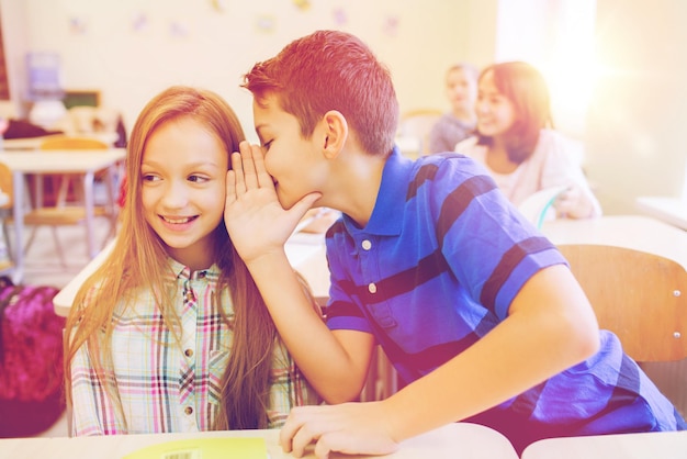 concetto di istruzione, scuola elementare, apprendimento e persone - scolaro sorridente che sussurra il segreto all'orecchio del compagno di classe in classe