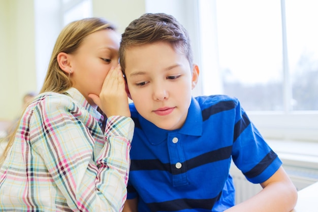 concetto di istruzione, scuola elementare, apprendimento e persone - studentessa sorridente che sussurra il segreto all'orecchio del compagno di classe in classe