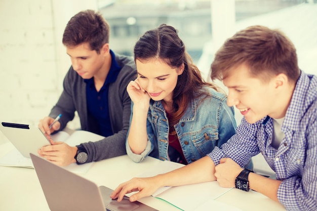 concetto di istruzione, tecnologia, scuola e internet - tre studenti sorridenti con laptop, tablet pc e notebook a scuola