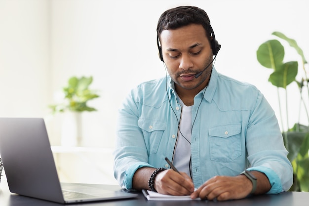 Concetto di lavoro a distanza. Giovane lavoratore di supporto indiano o libero professionista utilizza laptop e cuffie per comunicare con il cliente, prende appunti su un notebook, apprendimento online