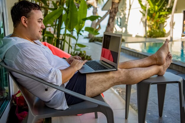 Concetto di lavoro a distanza uomo con laptop seduto a bordo piscina in un paese tropicale