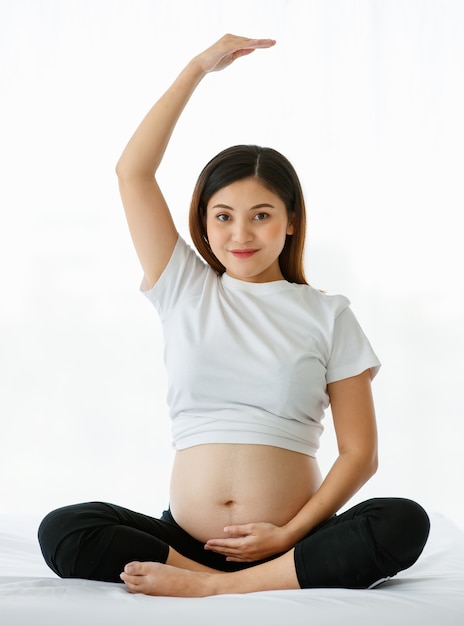 Concetto di madre sana. La giovane bella donna incinta che indossa una maglietta bianca e pantaloni neri seduta su un letto alza le mani sopra la testa facendo stretching dopo l'esercizio yoga.