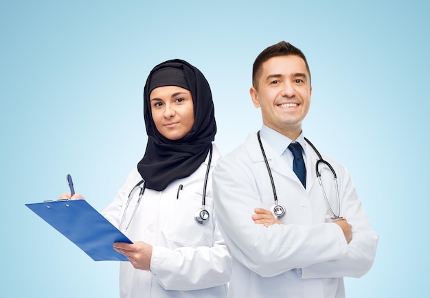 concetto di medicina, assistenza sanitaria e persone - medici sorridenti felici in camici bianchi con appunti e stetoscopi su sfondo blu