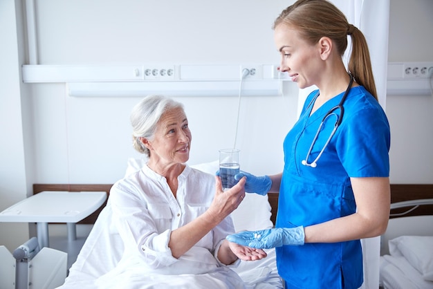 concetto di medicina, età, assistenza sanitaria e persone - infermiera che somministra farmaci e bicchiere d'acqua a una donna anziana nel reparto ospedaliero