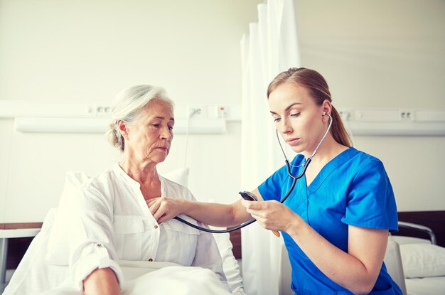 concetto di medicina, età, supporto, assistenza sanitaria e persone - medico o infermiere con stetoscopio in visita a una donna anziana e controllando il suo battito cardiaco nel reparto ospedaliero