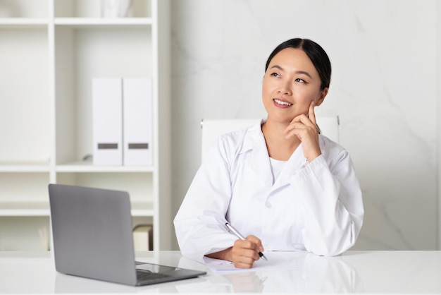 Concetto di medicina pensosa dottoressa asiatica seduta alla scrivania e che prende appunti