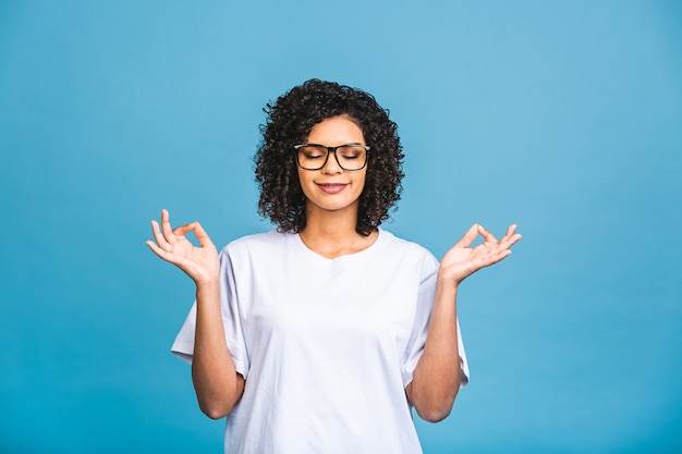 Concetto di meditazione. Bella giovane donna afroamericana si trova in posa meditativa, gode di un'atmosfera tranquilla, tiene le mani in gesto di preghiera, isolato su sfondo blu.