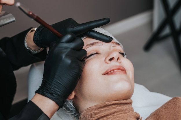 Concetto di microblading delle sopracciglia. Cosmetologo che prepara giovane donna per la procedura di trucco permanente del sopracciglio.