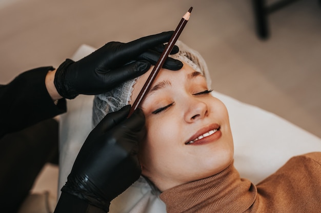 Concetto di microblading delle sopracciglia. Cosmetologo che prepara giovane donna per la procedura di trucco permanente del sopracciglio.