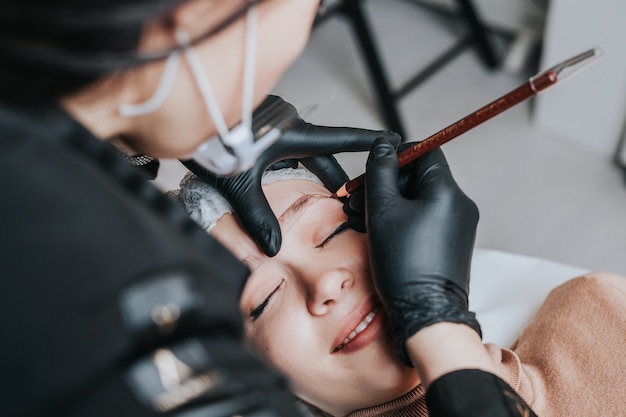 Concetto di microblading delle sopracciglia, procedura di trucco permanente delle sopracciglia. L'esperto di bellezza indossa una maschera facciale protettiva a causa della pandemia di Coronavirus.