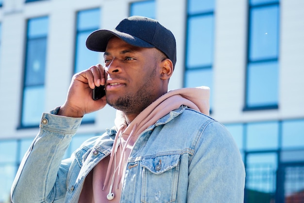 Concetto di moda di strada - uomo afroamericano elegante e bello in una camicia rosa con cappuccio e giacca di jeans e berretto nero contro le finestre del business center