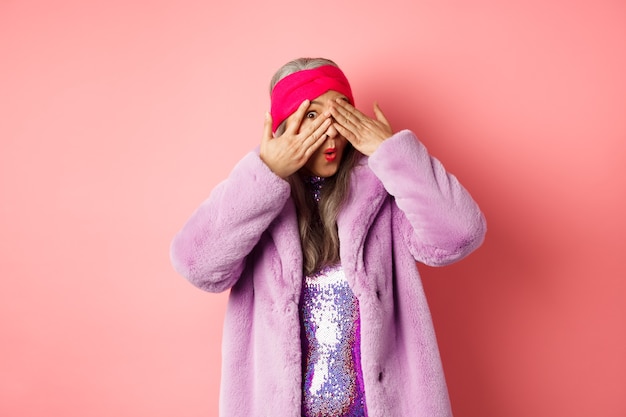 Concetto di moda e shopping. Elegante nonna hipster asiatica in abito viola che si copre gli occhi, sbirciando tra le dita con una faccia curiosa, spiando qualcosa di interessante, sfondo rosa.