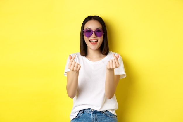 Concetto di moda e stile di vita. Attraente donna asiatica in occhiali da sole a forma di cuore, mostrando i cuori delle dita e sorridendo felice alla telecamera, in piedi su sfondo giallo.