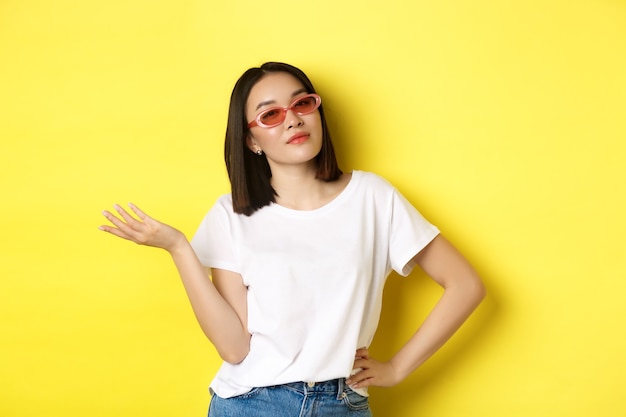 Concetto di moda e stile di vita. Donna asiatica sfacciata e sicura di sé con occhiali da sole alla moda che sembra sicura di sé alla telecamera, in piedi su sfondo giallo.