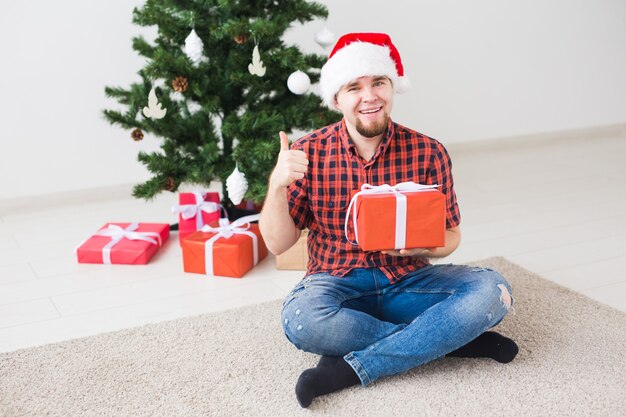 Concetto di Natale e festività - Uomo divertente con cappello da Babbo Natale che tiene un regalo a casa nel soggiorno.