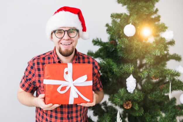 Concetto di Natale e festività - Uomo divertente con cappello da Babbo Natale che tiene un regalo a casa nel soggiorno.