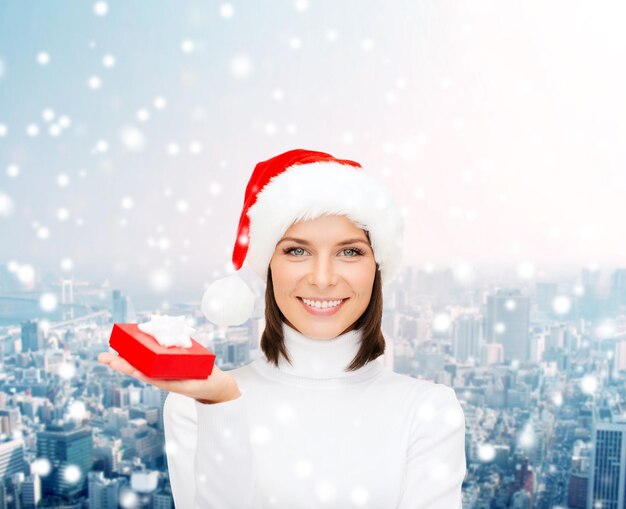 concetto di natale, inverno, felicità, vacanze e persone - donna sorridente con cappello da Babbo Natale con piccola confezione regalo rossa su sfondo di città innevata