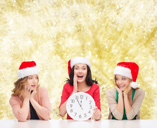 concetto di natale, inverno, vacanze, tempo e persone - donne sorridenti in cappelli di Babbo Natale con orologio su sfondo a luci gialle