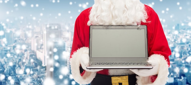 concetto di natale, pubblicità, tecnologia e persone - primo piano di Babbo Natale con un computer portatile su sfondo di città innevata
