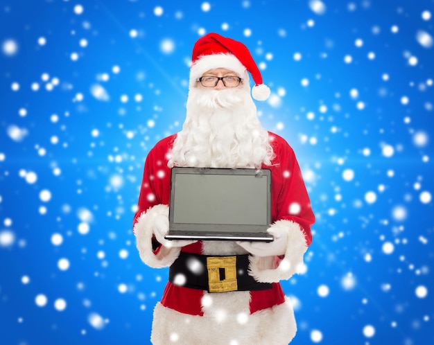 concetto di natale, pubblicità, tecnologia e persone - uomo in costume di babbo natale con computer portatile su sfondo blu innevato