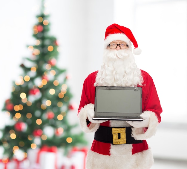 concetto di natale, pubblicità, tecnologia e persone - uomo in costume di babbo natale con computer portatile sul soggiorno con sfondo ad albero