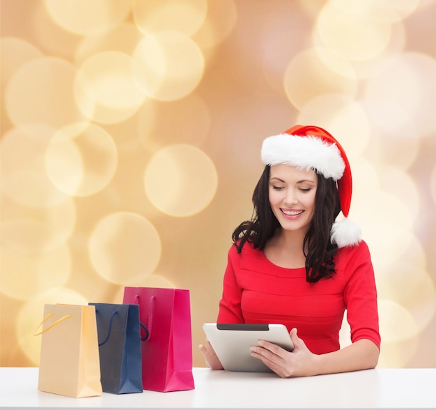 concetto di natale, tecnologia e persone - donna sorridente in cappello di Babbo Natale con borse della spesa e computer tablet pc su sfondo di luci beige