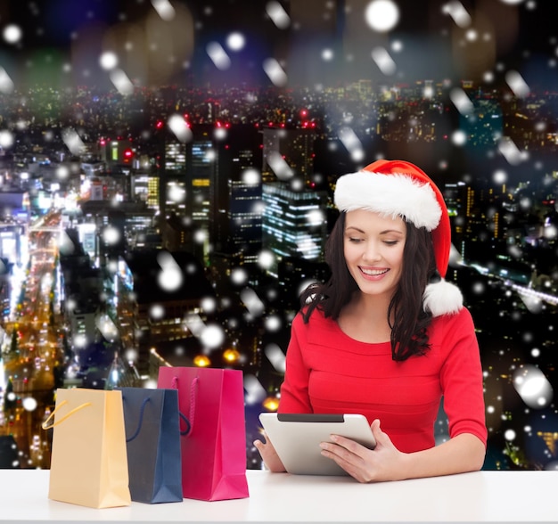 concetto di natale, tecnologia e persone - donna sorridente in cappello di Babbo Natale con borse della spesa e computer tablet pc su sfondo nevoso della città di notte