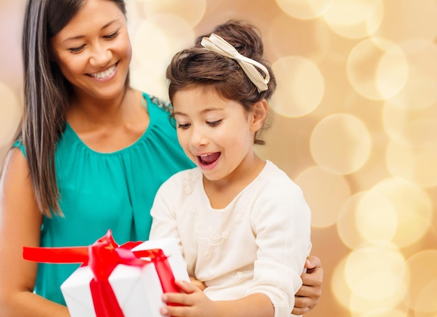 concetto di natale, vacanze, celebrazione, famiglia e persone - madre felice e bambina con confezione regalo su sfondo di luci beige