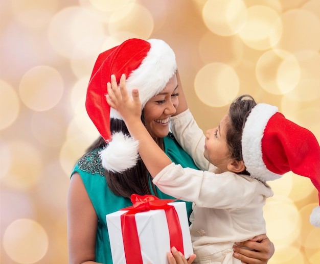 concetto di natale, vacanze, celebrazione, famiglia e persone - madre felice e bambina in cappelli di Babbo Natale con confezione regalo su sfondo di luci beige