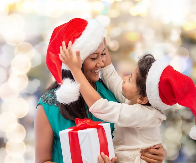 concetto di natale, vacanze, celebrazione, famiglia e persone - madre felice e bambina in cappelli di Babbo Natale con confezione regalo su sfondo di luci