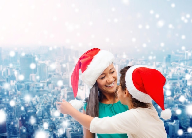 concetto di natale, vacanze, celebrazione, famiglia e persone - madre felice e bambina in cappelli di Babbo Natale su sfondo di città innevata