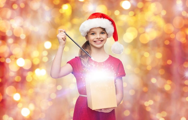 concetto di natale, vacanze e bambini - ragazza sorridente in cappello di santa helper con confezione regalo e bacchetta magica su sfondo di luci