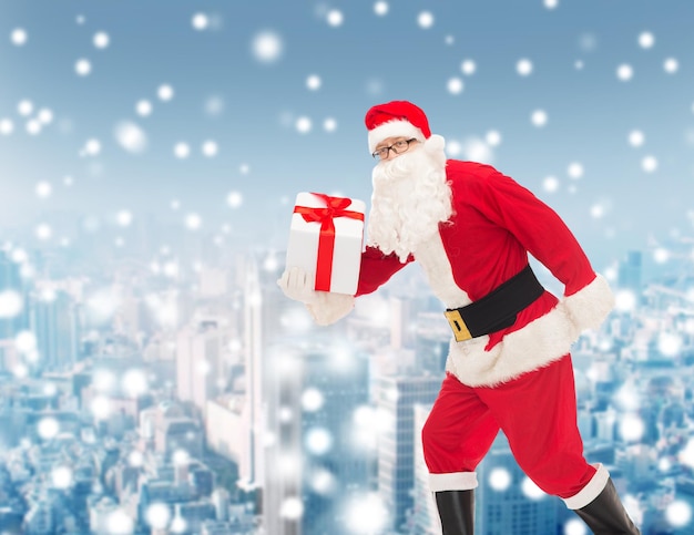 concetto di natale, vacanze e persone - uomo in costume di babbo natale che corre con confezione regalo su sfondo città innevata