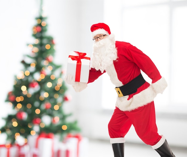 concetto di natale, vacanze e persone - uomo in costume di babbo natale che corre con confezione regalo su sfondo soggiorno e albero