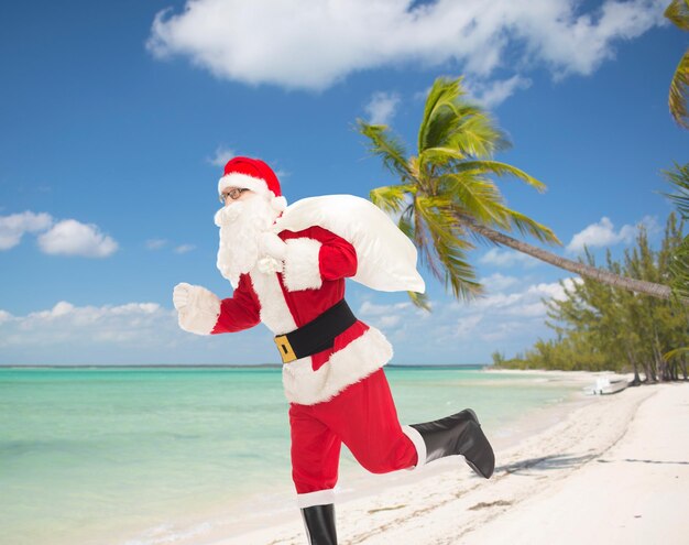 concetto di natale, vacanze e persone - uomo in costume di babbo natale che corre con la borsa su sfondo spiaggia tropicale