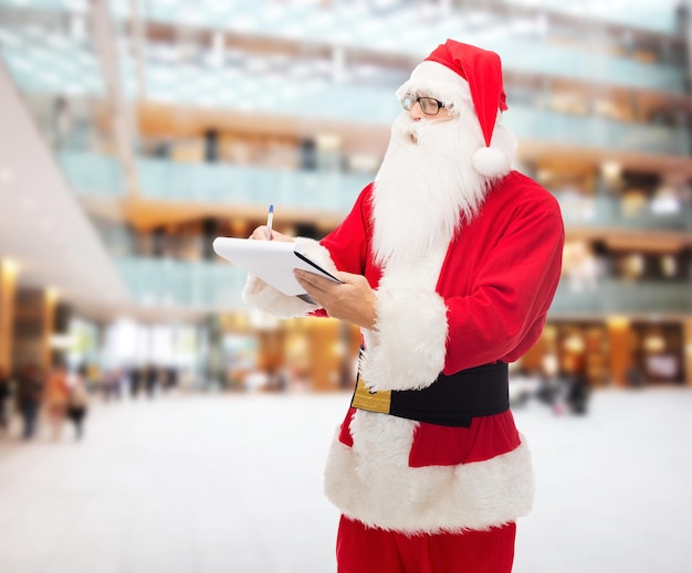 concetto di natale, vacanze e persone - uomo in costume di babbo natale con blocco note e penna su sfondo centro commerciale