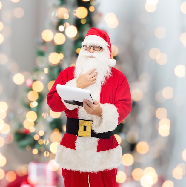 concetto di natale, vacanze e persone - uomo in costume di babbo natale con blocco note su sfondo di luci ad albero