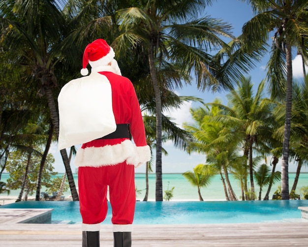 concetto di natale, vacanze e persone - uomo in costume di babbo natale con borsa dal retro su sfondo spiaggia tropicale e piscina