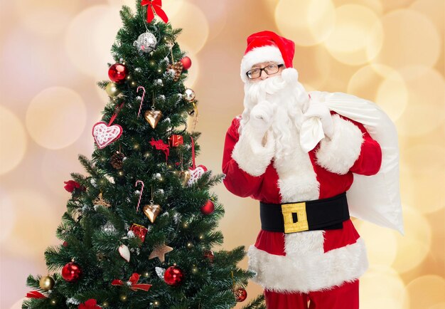 concetto di natale, vacanze e persone - uomo in costume di babbo natale con borsa e albero di natale che fa un gesto silenzioso su sfondo di luci beige