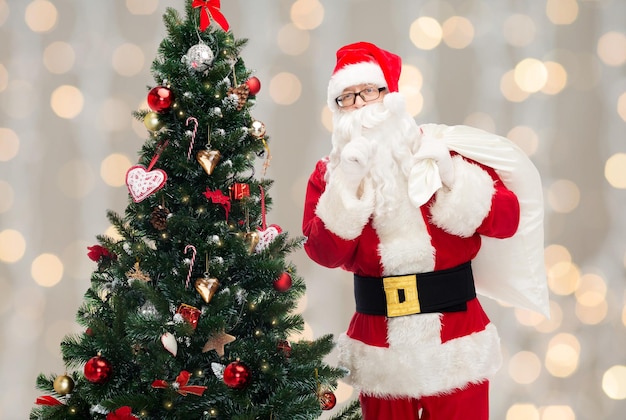 concetto di natale, vacanze e persone - uomo in costume di babbo natale con borsa e albero di natale che fa un gesto silenzioso su sfondo di luci