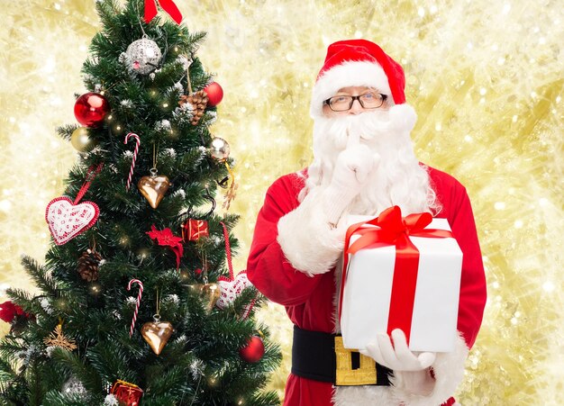 concetto di natale, vacanze e persone - uomo in costume di babbo natale con confezione regalo e albero che fa un gesto silenzioso su sfondo a luci gialle