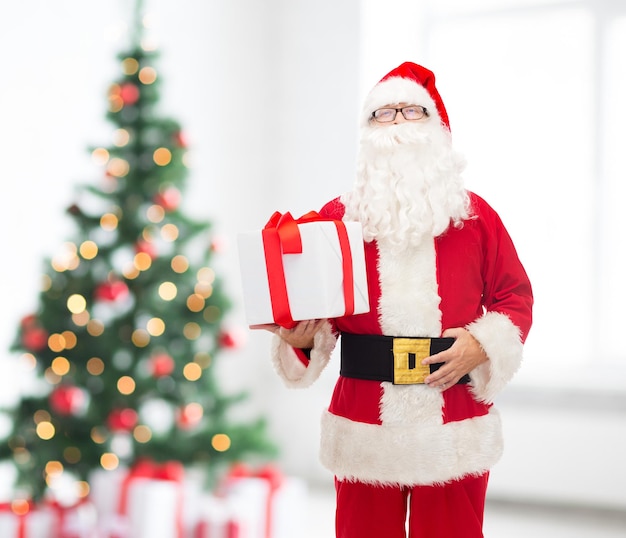 concetto di natale, vacanze e persone - uomo in costume di babbo natale con confezione regalo su sfondo soggiorno e albero